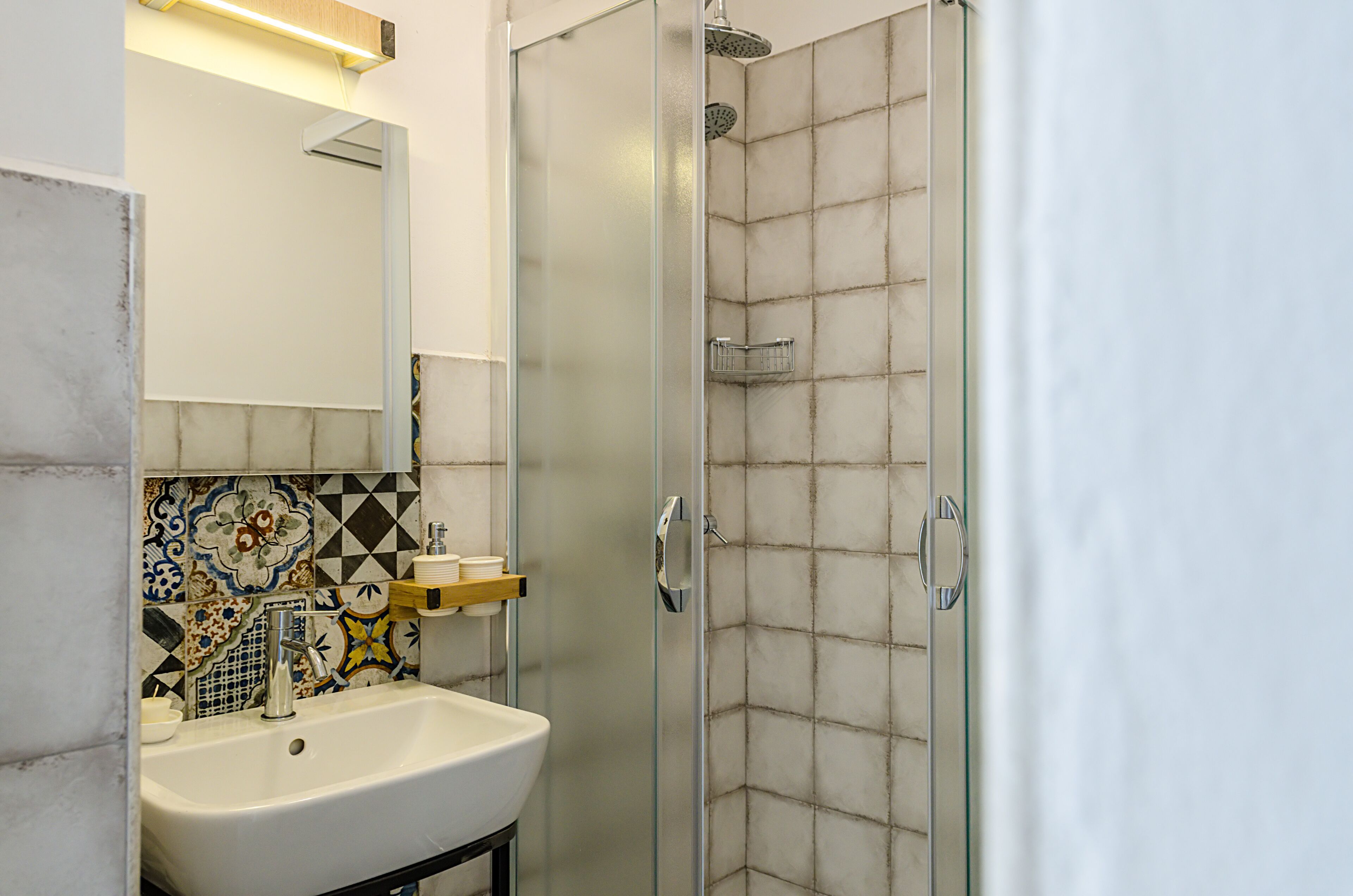 Un baño con ducha de vidrio, lavamanos blanco y salpicadero de azulejos con diversos diseños.