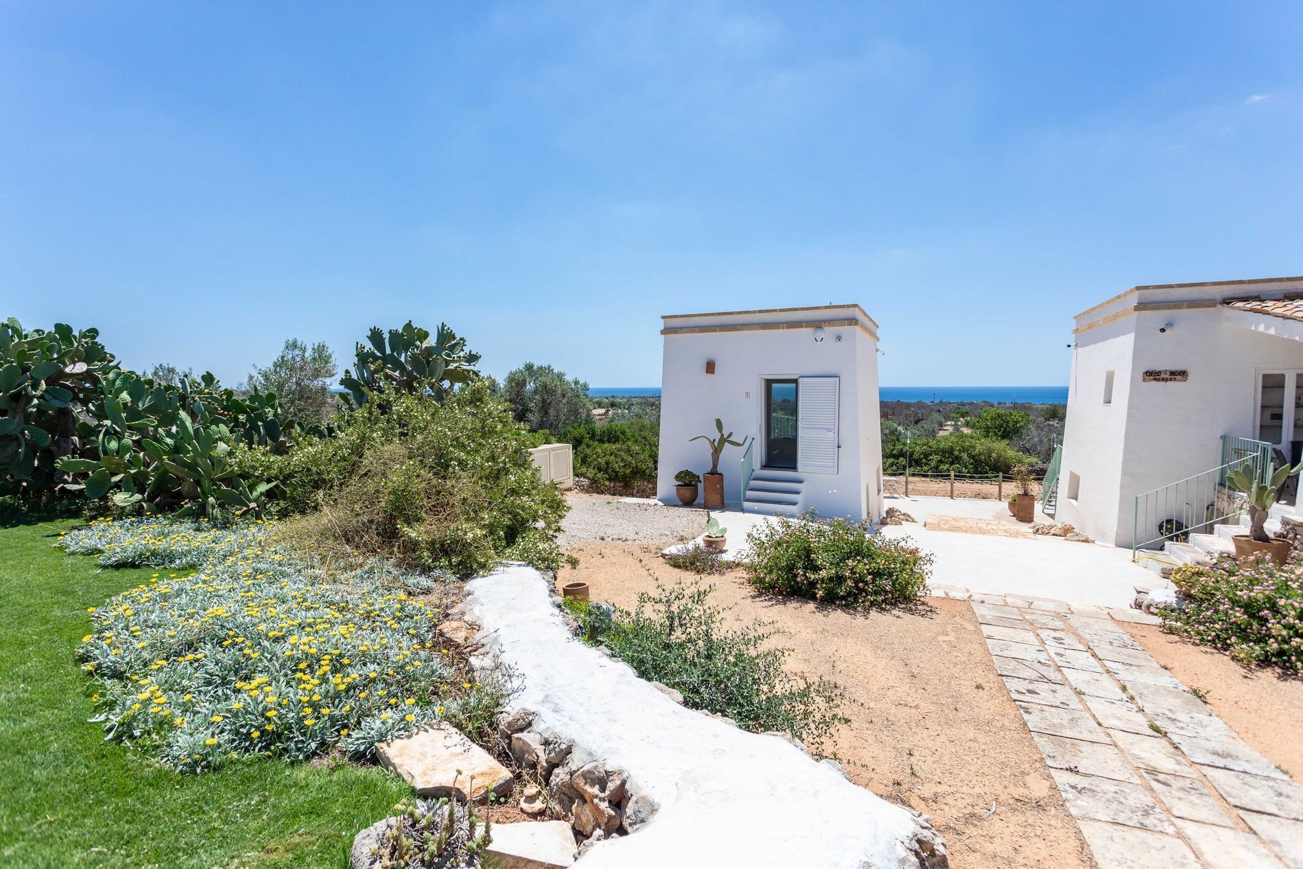 Una propiedad costera con dos edificios blancos pequeños, un jardín con flores amarillas y una vista despejada al mar.