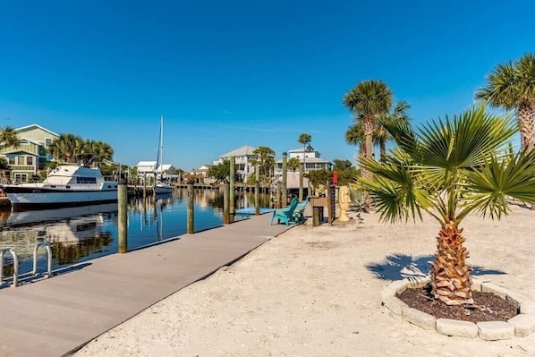 A marina with boats docked, a sandy walkway, and palm trees.