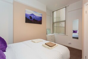 A bedroom with a bed, a nightstand, a window with blinds, a painting on the wall, and a mirror on the wall. - Cherry Property - Passionfruit Suite (Blackpool)