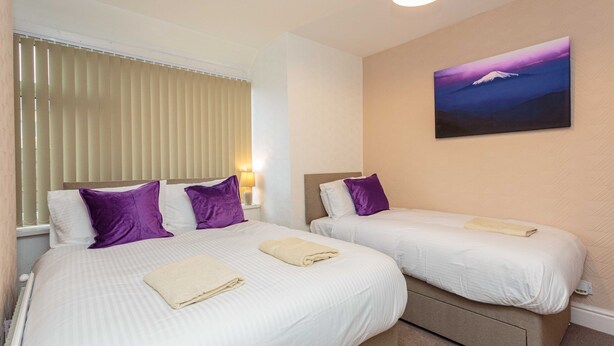 Hotellihuoneessa on kaksi sänkyä, joissa on valkoiset lakanat ja violetit tyynyt. Seinällä on taulu, ikkunassa sälekaihtimet ja yöpöydällä lamppu.