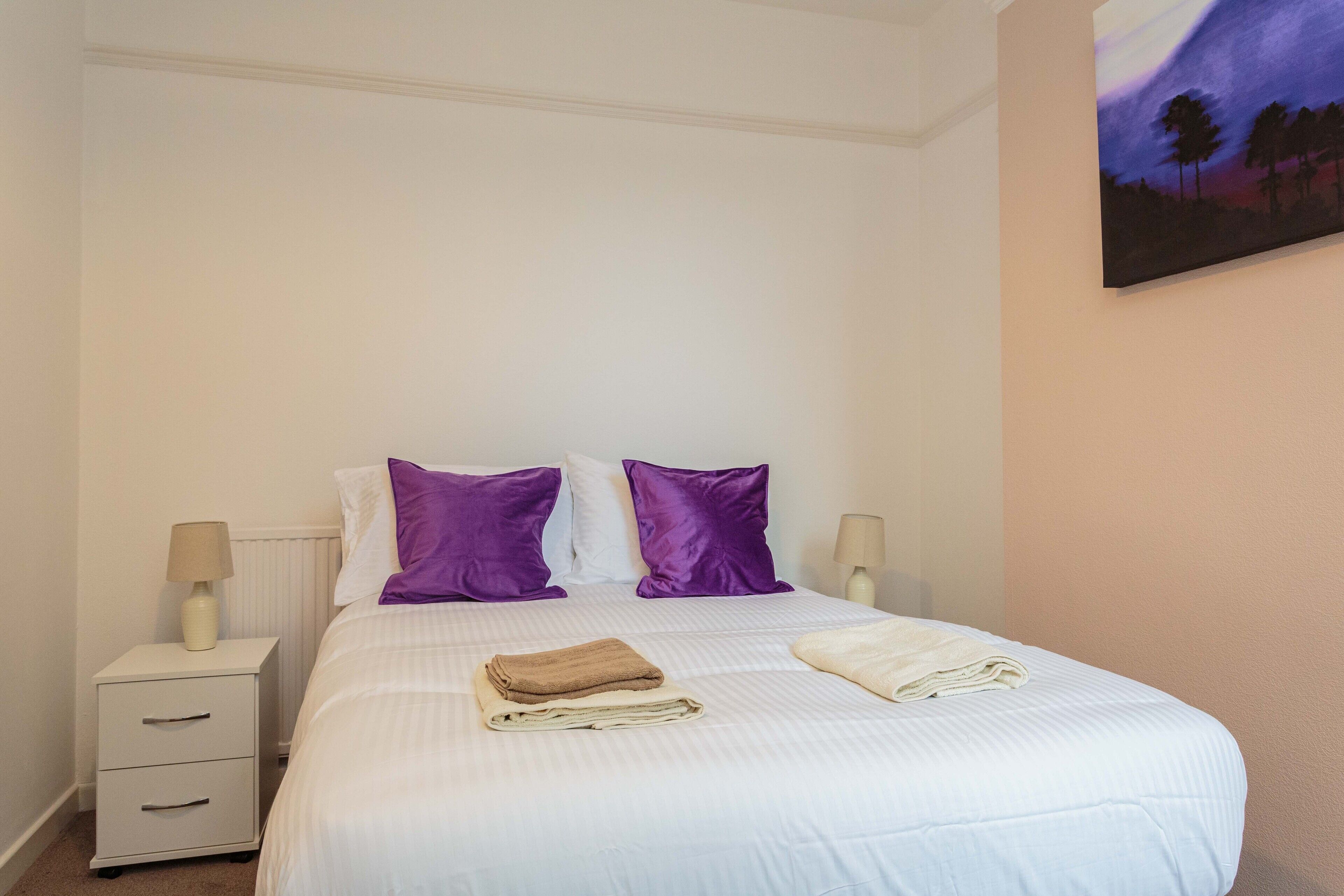A neatly made bed with white and purple bedding, a small bedside table with a lamp, and a painting on the wall.