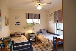 Une chambre avec deux lits simples, un ventilateur de plafond, une fenêtre avec des rideaux à rayures et une petite table.