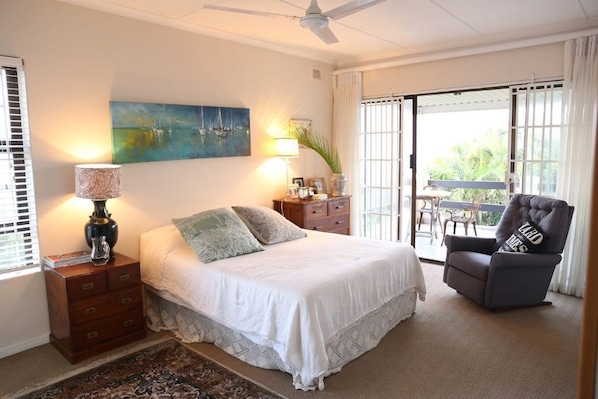 Une chambre à coucher comprenant un lit, une table de chevet, une chaise, un tableau et une vue sur l’extérieur, visible à travers une porte coulissante.
