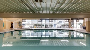 Une piscine intérieure avec des parois en verre transparent, un plafond en métal et une vue sur les bâtiments environnants.