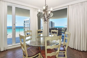 A dining area with a glass table and wooden chairs, large windows with curtains, and a view of the beach and buildings.