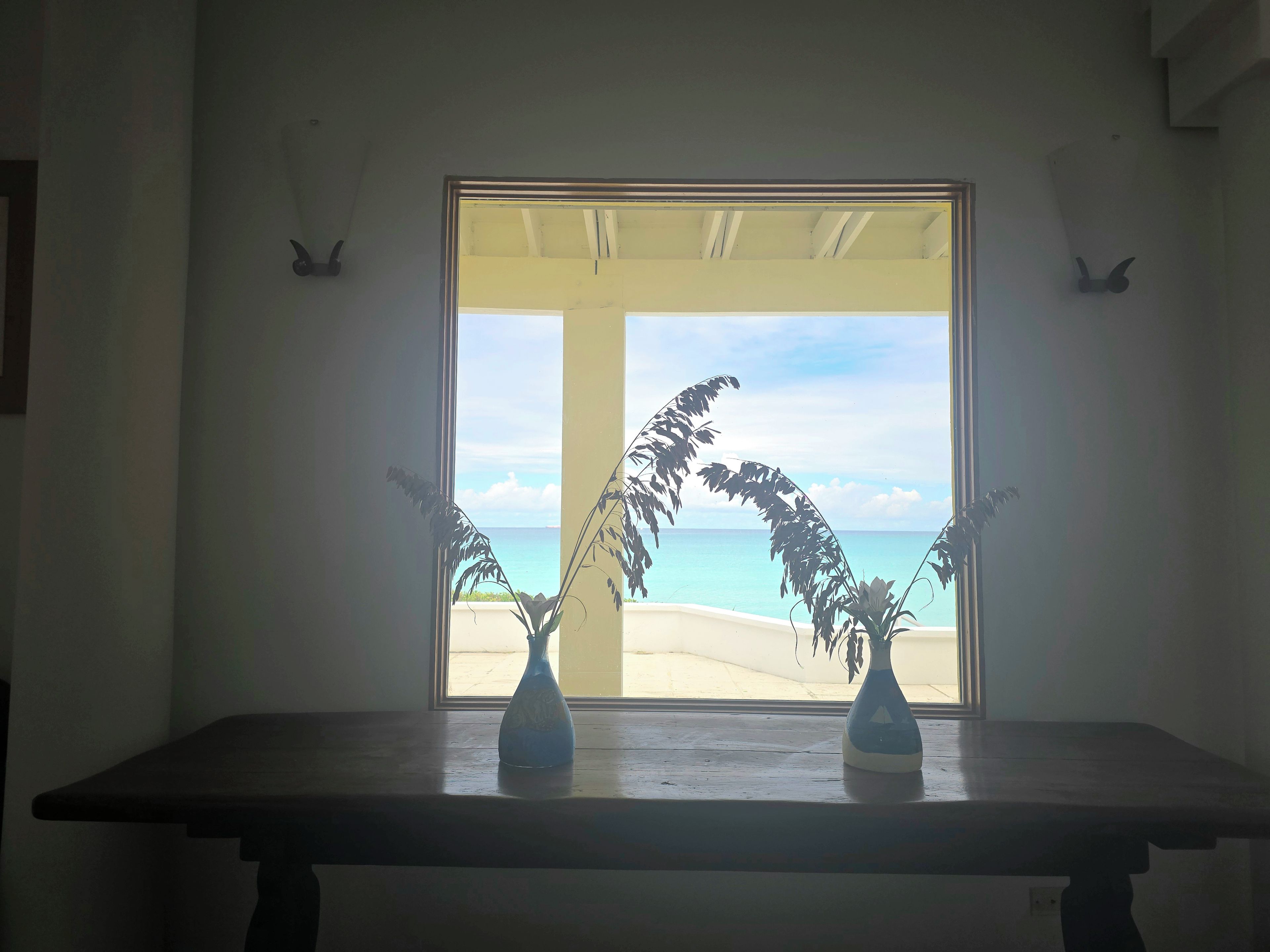 Une pièce dotée d’une grande fenêtre donnant sur une plage, d’une table en bois et de deux vases posés sur la table.