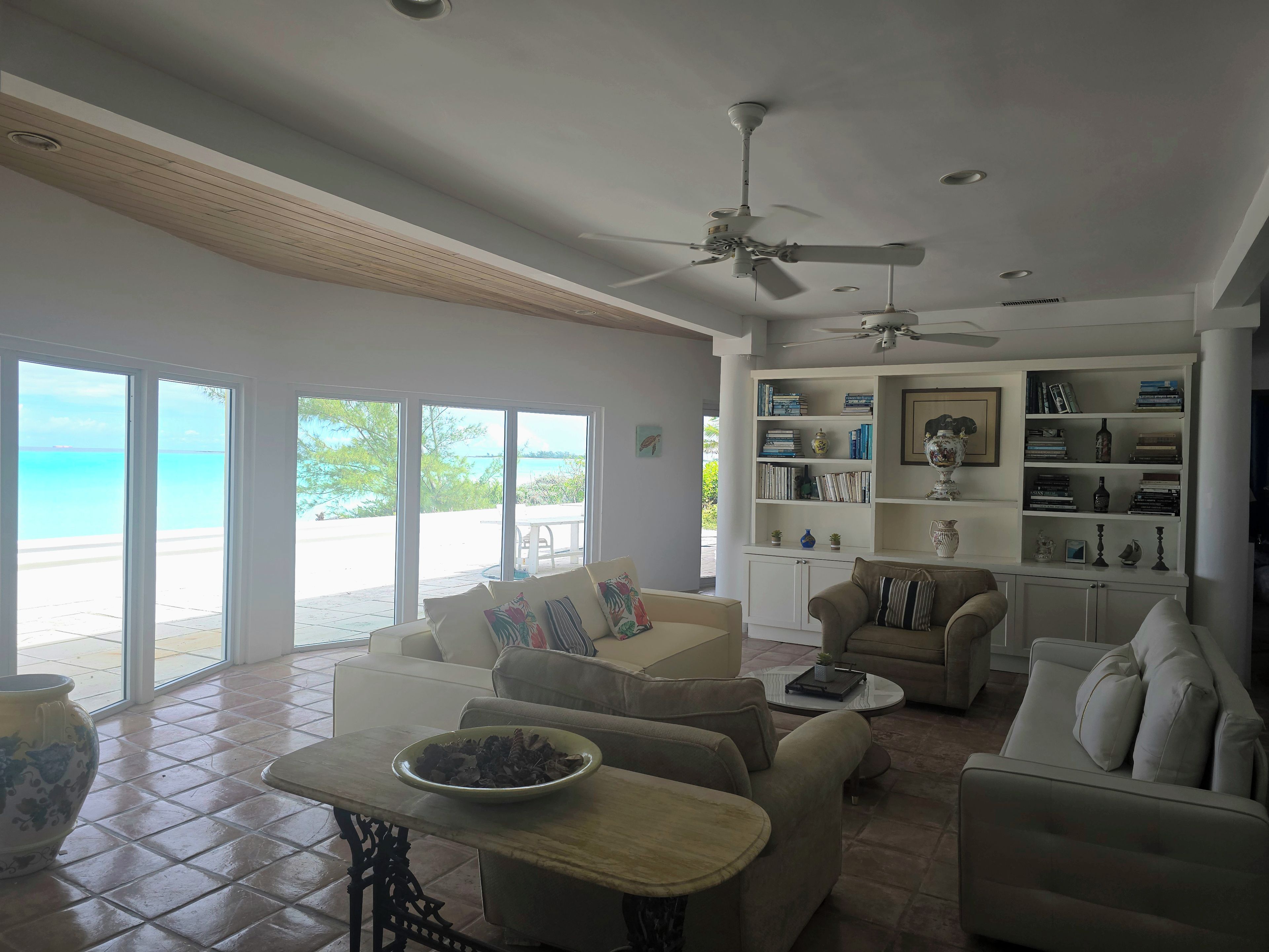 Un salon spacieux doté d’une grande fenêtre donnant sur une plage, d’un ventilateur de plafond et d’une biblioth èque remplie de livres et d’objets décoratifs.