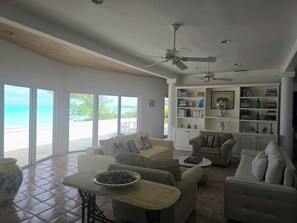 A spacious living room with a large window overlooking a beach, a ceiling fan, and a bookshelf filled with books and decorative items.