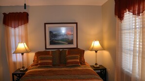 A neatly made bed with striped bedding, two bedside lamps, a framed landscape painting above the bed, and sheer curtains with a draped valance.
