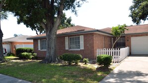 A single-story brick house with a white picket fence, a driveway, and a large tree in the front yard.
