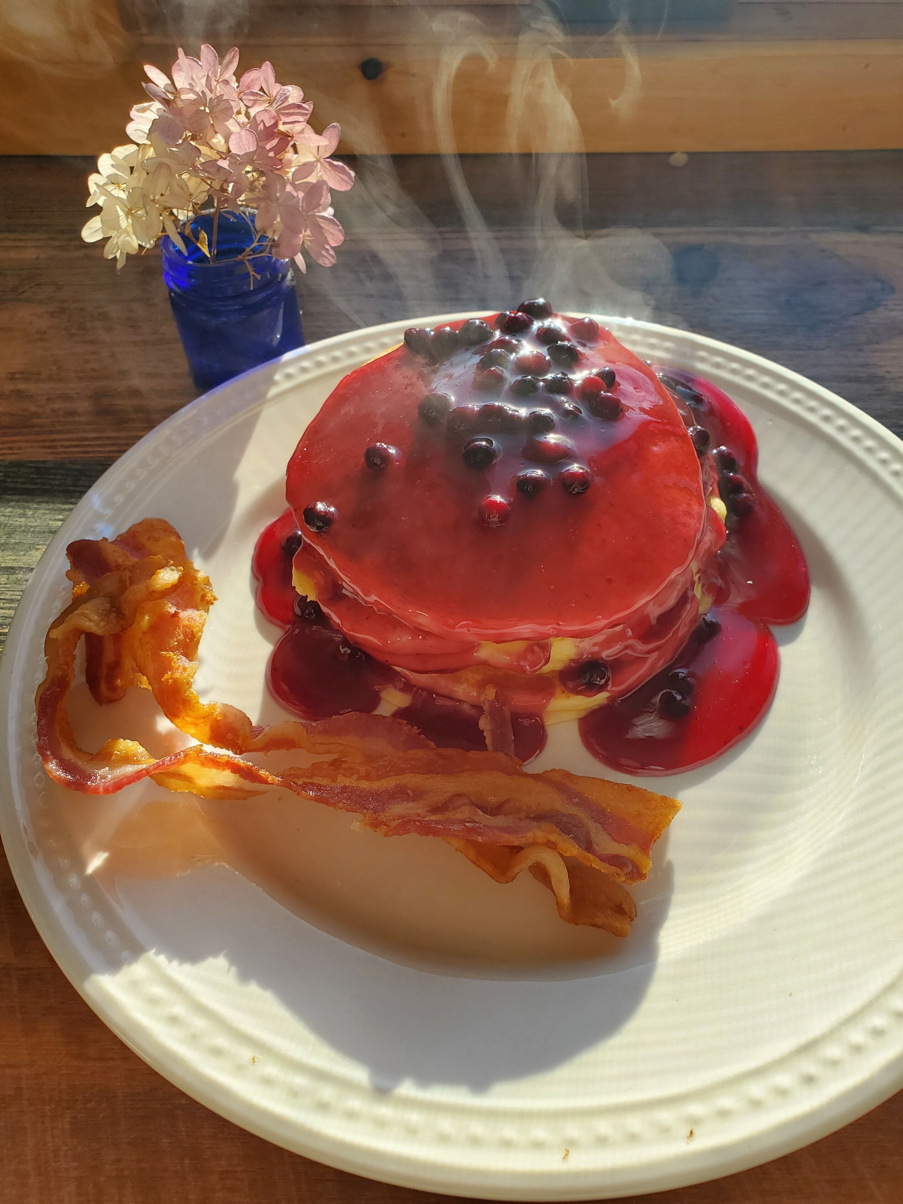A plate with pancakes topped with blueberries and syrup, accompanied by bacon strips and a small blue vase with flowers.