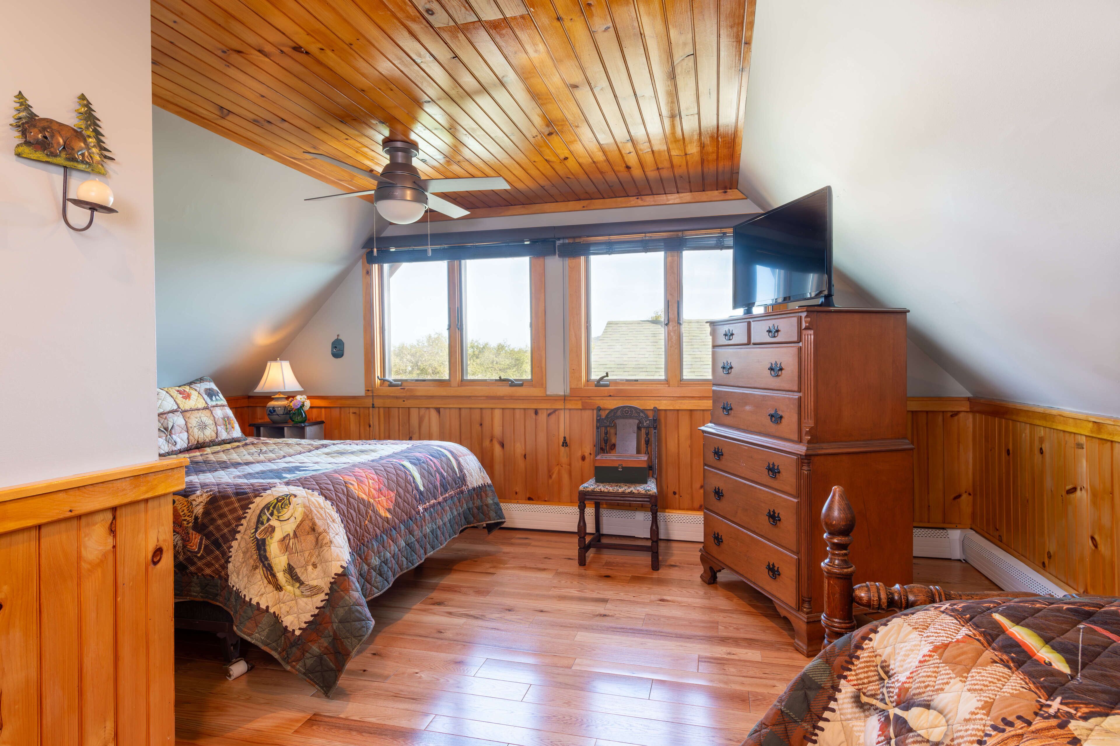 A bedroom with a wooden ceiling, a bed with a quilt, a dresser, a chair, and a window with a view of trees.