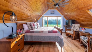 Une chambre avec un plafond en bois, un lit recouvert d’une courtepointe, une table de chevet, une lampe, une chaise et un téléviseur.