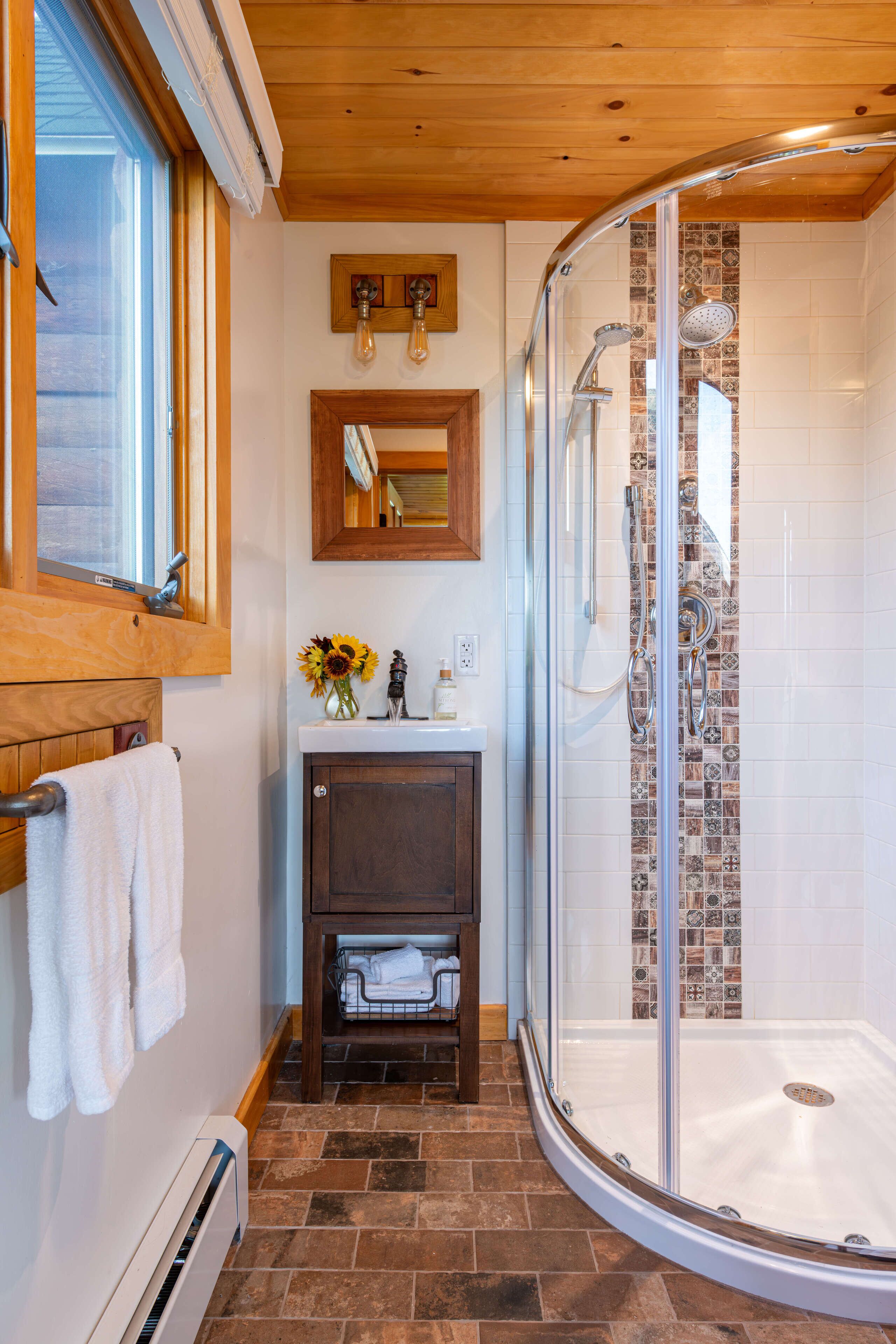 A bathroom with a glass shower enclosure, a wooden vanity, a sink with a mirror, and a window.