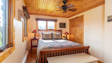 Quarto com teto de madeira, cama com colcha, duas luminárias de cabeceira e janela com persianas.