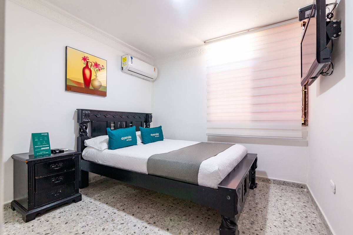 A bedroom with a bed, a nightstand, a painting, an air conditioning unit, and a window with blinds.