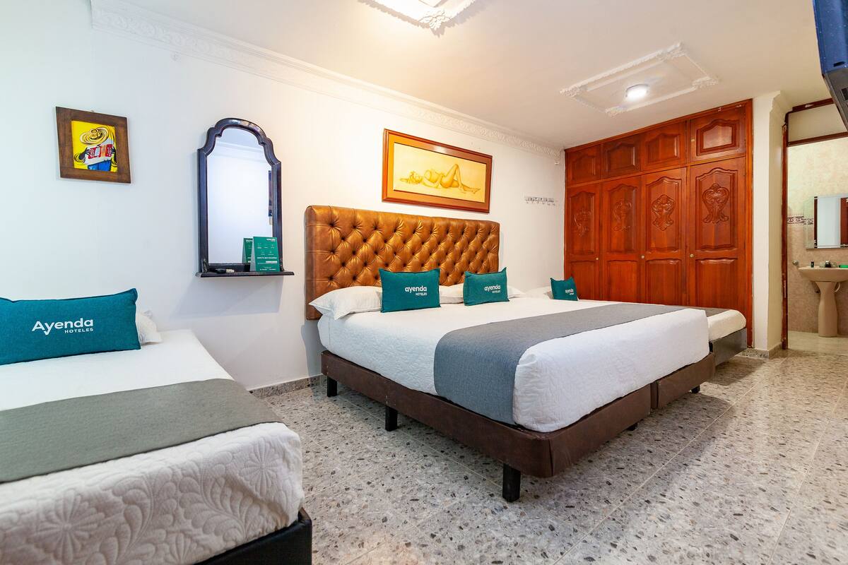 A hotel room with a large bed, two smaller beds, a wooden wardrobe, a mirror, and artwork on the walls.