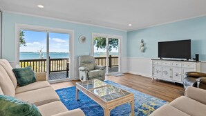 A living room with a large glass coffee table, beige sofas, a TV on a white cabinet, and a view of the ocean through large sliding glass doors. - Bogue Sound Cottage with Boat Dock & Pet Friendly (Emerald Isle)