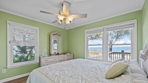 A bedroom with a large bed, a dresser, a ceiling fan, and a view of the ocean through the windows. - Bogue Sound Cottage with Boat Dock & Pet Friendly (Emerald Isle)