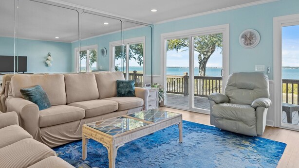 A living room with a beige sofa, a glass coffee table, and a view of the ocean through large sliding glass doors. - Bogue Sound Cottage with Boat Dock & Pet Friendly (Emerald Isle)
