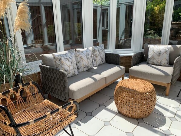 En solbeskinnet terrasse med en sofa, to lænestole, en flettet bænk og en potteplante.
