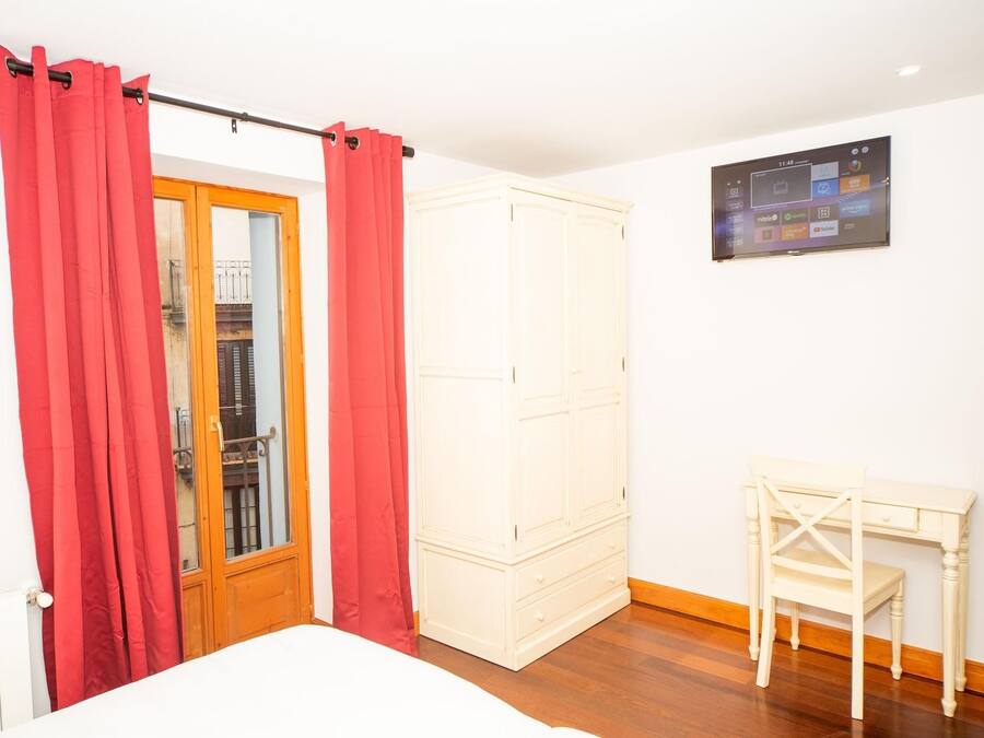 A bedroom with a bed, a wardrobe, a desk with a chair, and a TV on the wall.