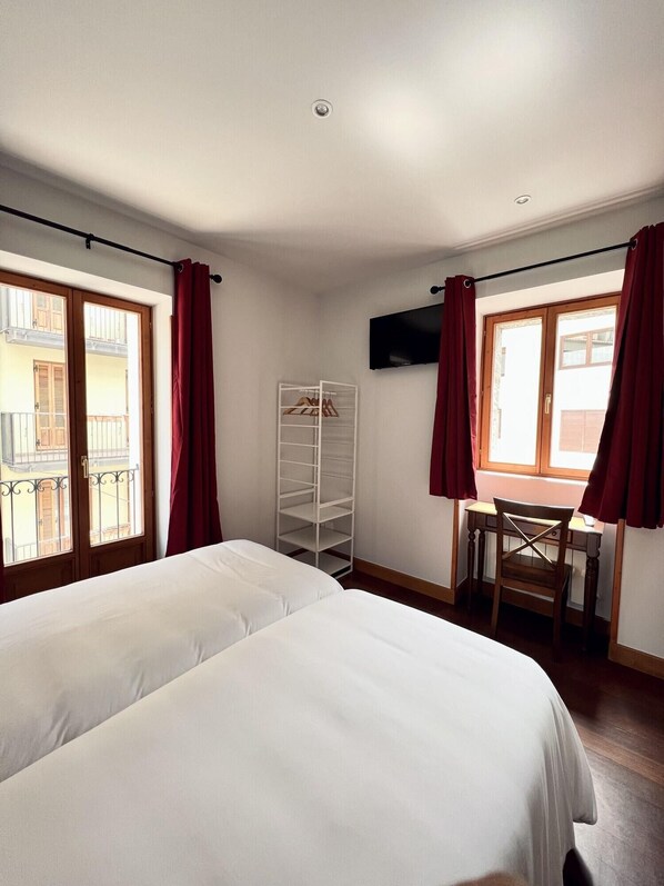 A hotel room with two beds, a desk, a chair, a TV mounted on the wall, and a window with red curtains. - Hotel Branka (Mundaka)