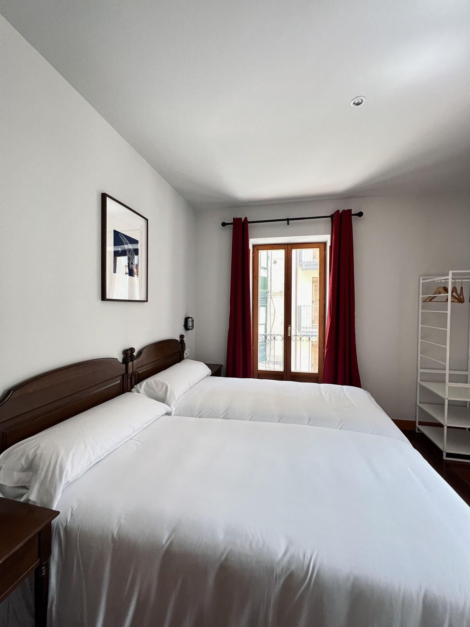 Un dormitorio con dos camas, una ventana con cortinas rojas, un armario blanco y un cuadro en la pared.