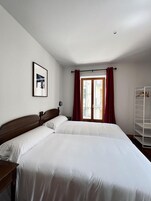 Un dormitorio con dos camas, una ventana con cortinas rojas, un armario blanco y un cuadro en la pared.