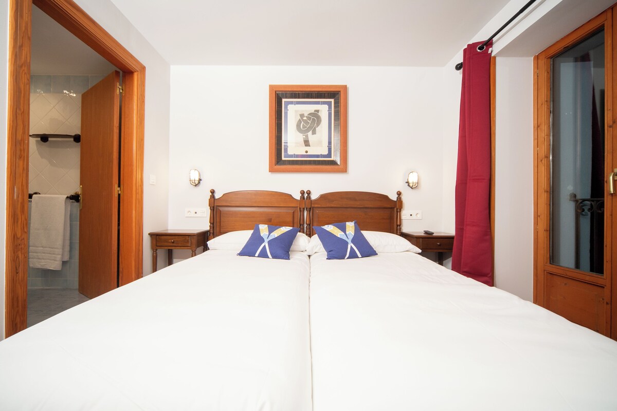 A double bed with a wooden headboard, two bedside tables, a framed picture on the wall, and a red curtain on the window.