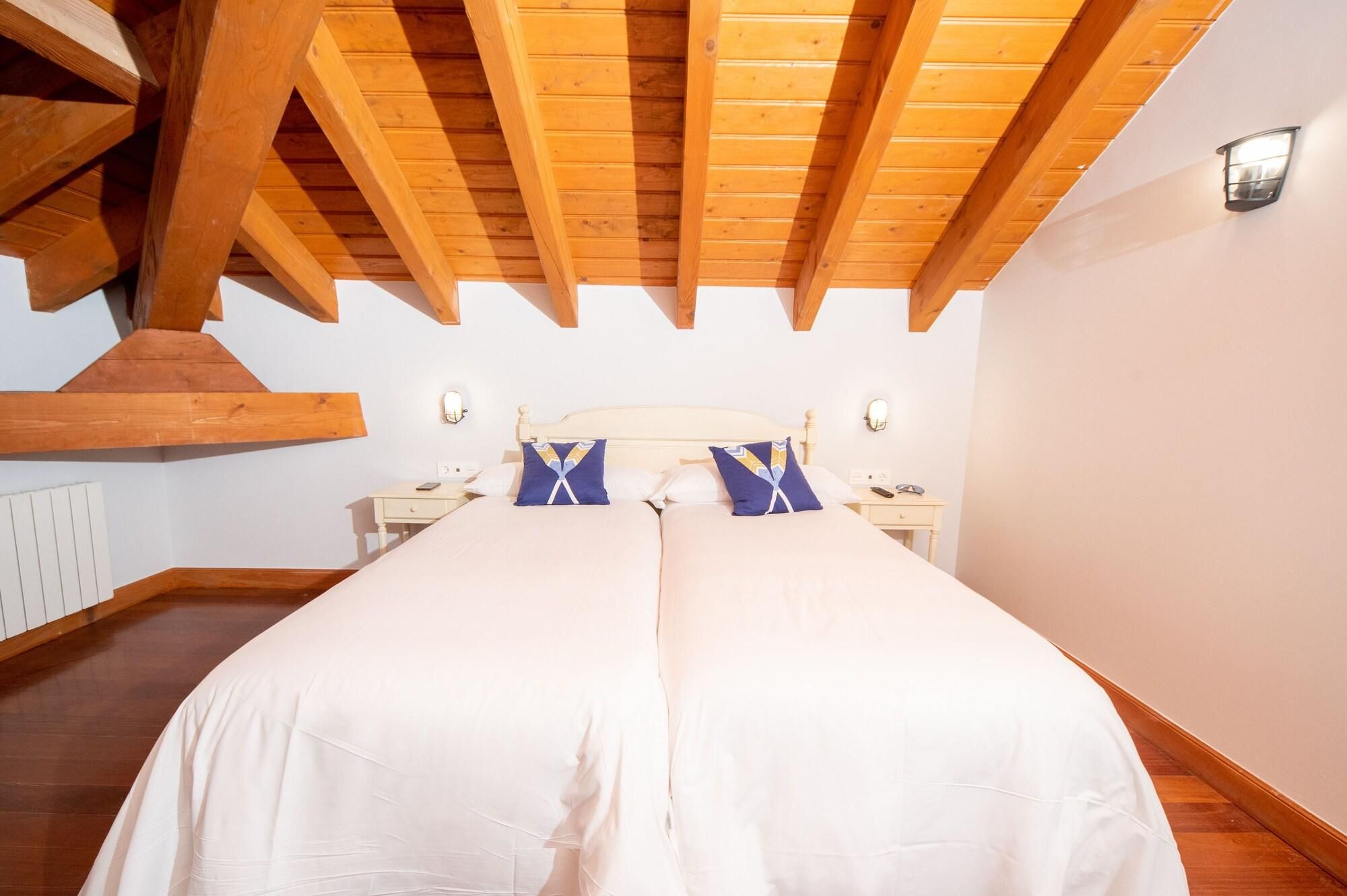 Un dormitorio con una cama grande, vigas de madera, paredes blancas y una mesita de noche con lámpara.