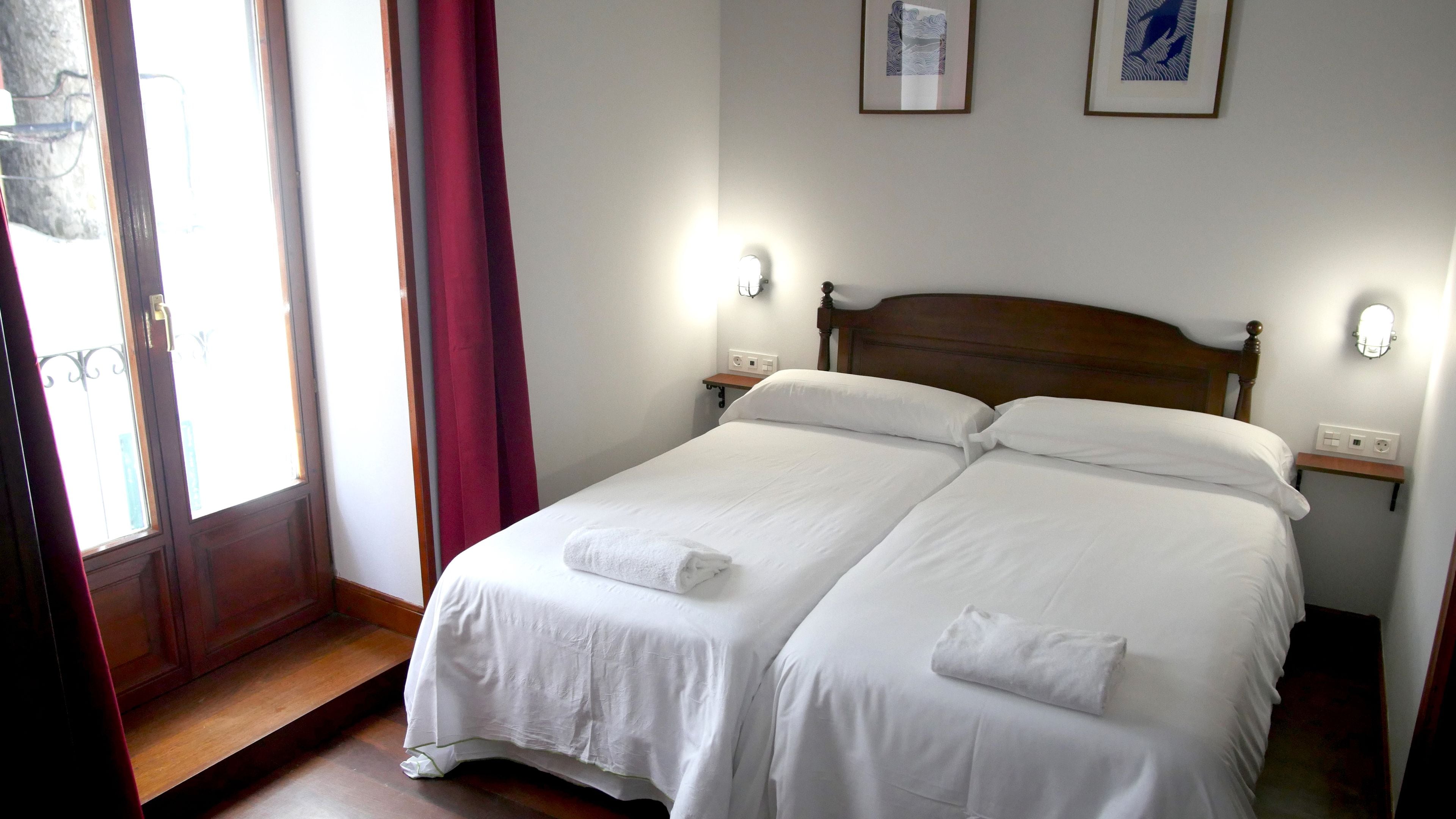 Cama doble con sábanas blancas y dos toallas, cabecera de madera, mesitas de noche con lámparas y dos cuadros en la pared.