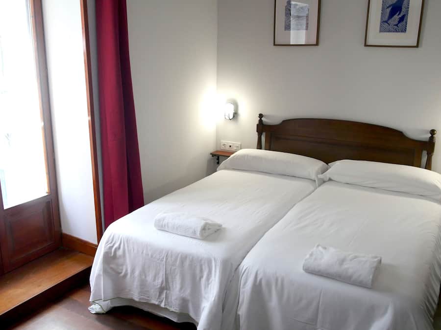 Een tweepersoonsbed met witte lakens en twee handdoeken, een houten hoofdbord, nachtkastjes met lampen en twee ingelijste foto’s aan de muur.