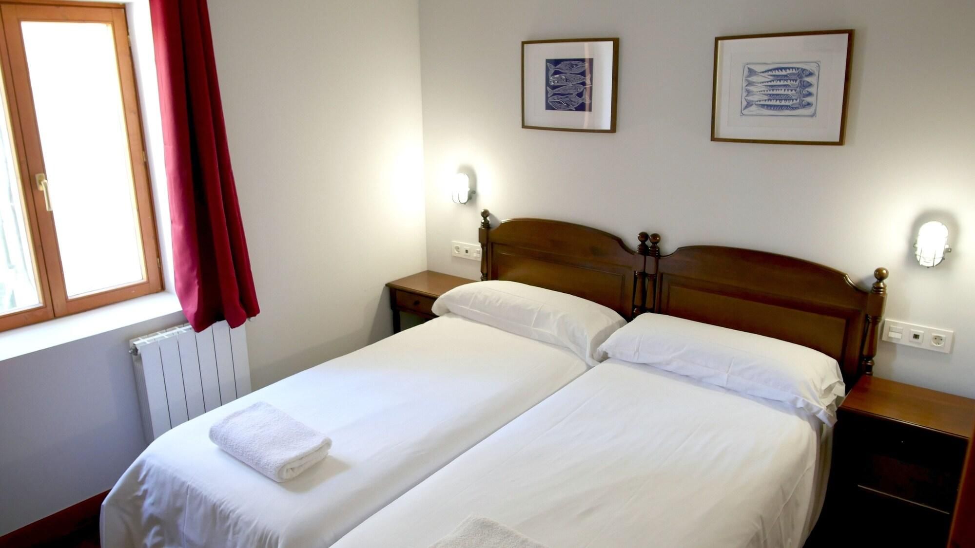 Una cama doble con sábanas blancas, dos mesitas de noche, una ventana con cortinas rojas y dos cuadros en la pared.