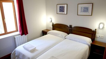 Una cama doble con sábanas blancas, dos mesitas de noche, una ventana con cortinas rojas y dos cuadros en la pared.