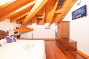 A bedroom with a sloped ceiling, wooden beams, a bed with pillows, a desk with a laptop, a wardrobe, and an air conditioning unit. - Hotel Branka (Mundaka)