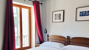 A hotel room with two beds, a wooden headboard, a nightstand, a wall-mounted lamp, and a window with red curtains. - Hotel Branka (Mundaka)