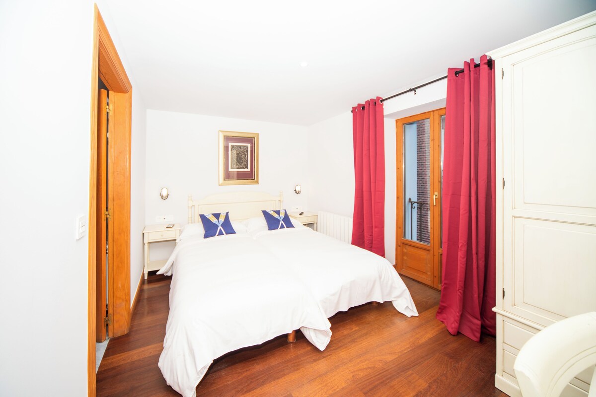 A bedroom with a wooden floor, a bed with white bedding, a nightstand, a bedside table, a framed picture on the wall, and a door with red curtains.