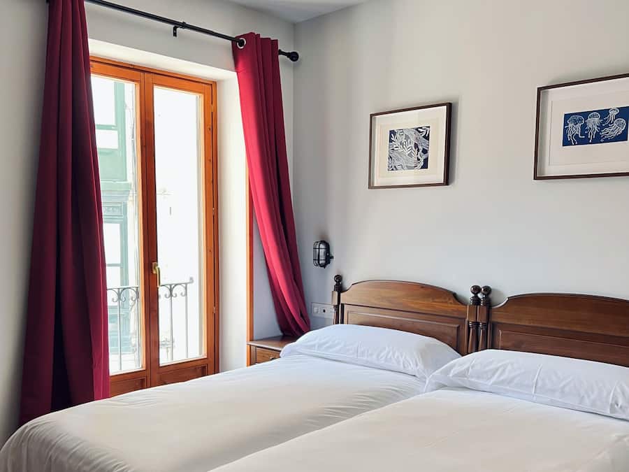 A hotel room with two beds, a wooden headboard, a nightstand, a wall-mounted lamp, and a window with red curtains.