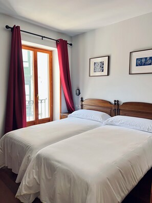 A hotel room with two beds, a wooden headboard, a nightstand, a wall-mounted lamp, and a window with red curtains. - Hotel Branka (Mundaka)