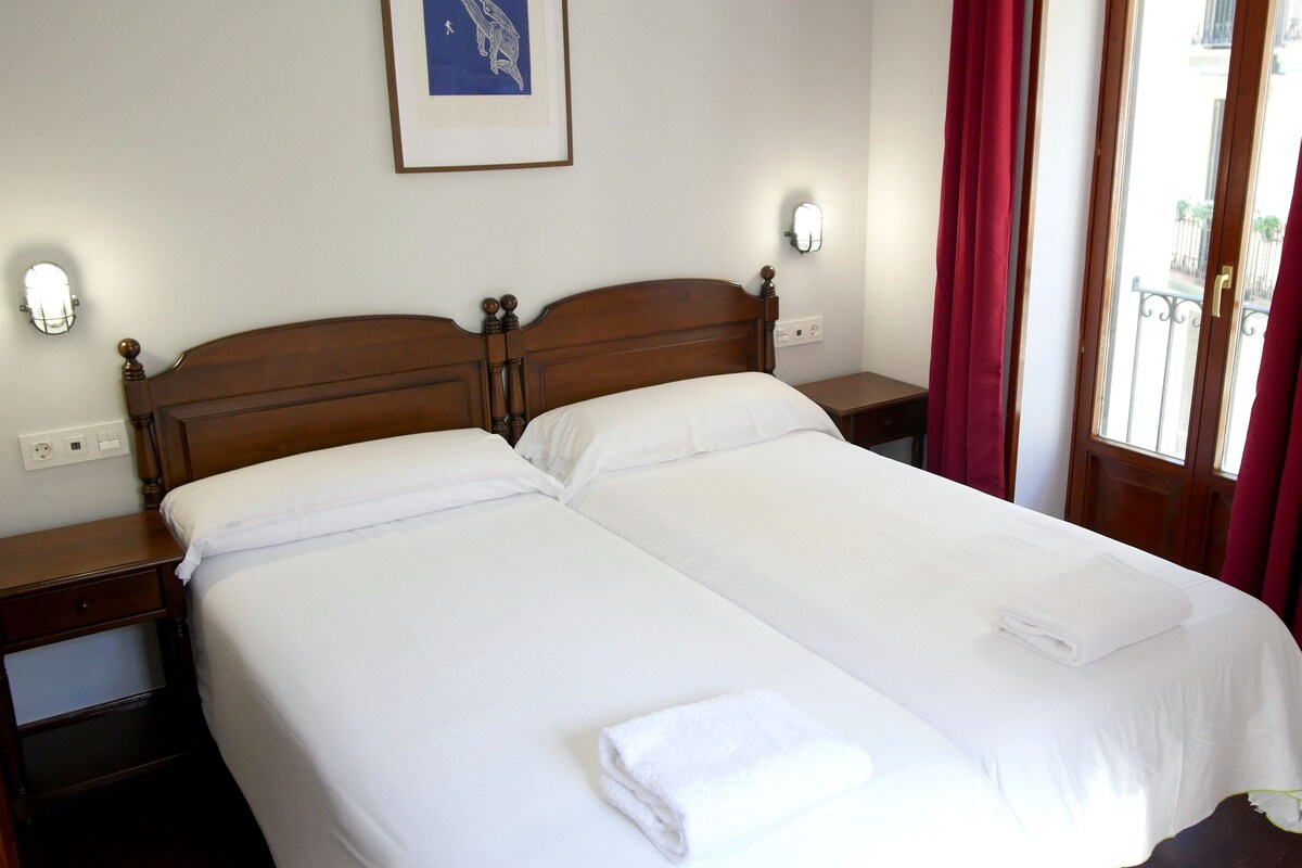 A double bed with white linens and a folded towel, wooden headboard, bedside tables with lamps, a framed artwork on the wall, and red curtains.