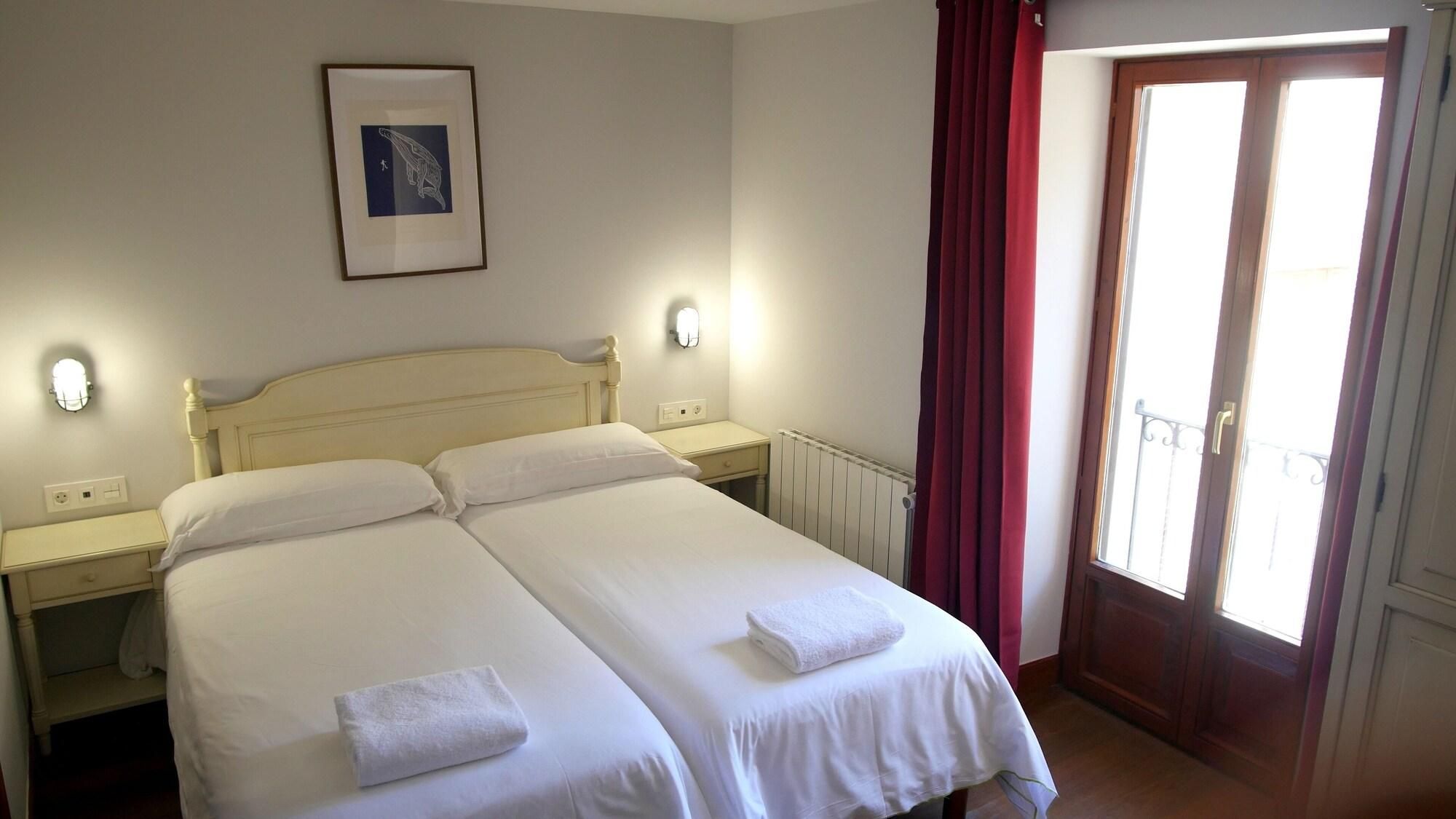 Cama doble con dos almohadas y dos toallas, una mesita de noche, una lámpara de pared, un cuadro enmarco, una ventana con cortinas rojas y un radiador.
