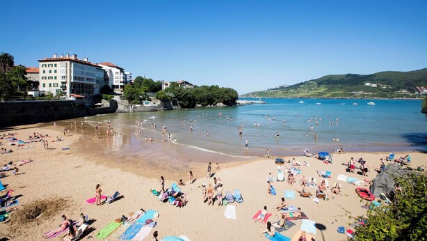 A crowded beach with people sunbathing and swimming, buildings in the background, and boats in the water. - Hotel Branka (Mundaka)
