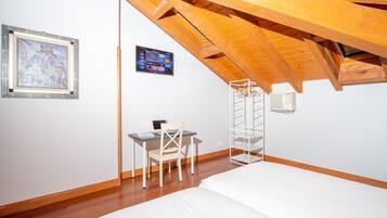 Un dormitorio con techo inclinado, una cama, un escritorio con silla, un televisor montado en la pared y un cuadro enmarco.