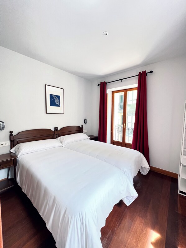 A double bed with white bedding, a wooden headboard, a nightstand, a wall-mounted lamp, and a window with red curtains. - Hotel Branka (Mundaka)