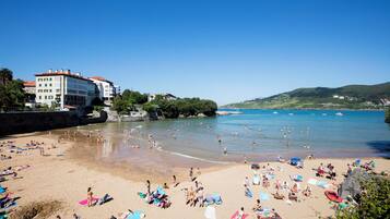 Una playa concurrida, gente tomando el sol y nadando, edificios al fondo y barcos en el agua.