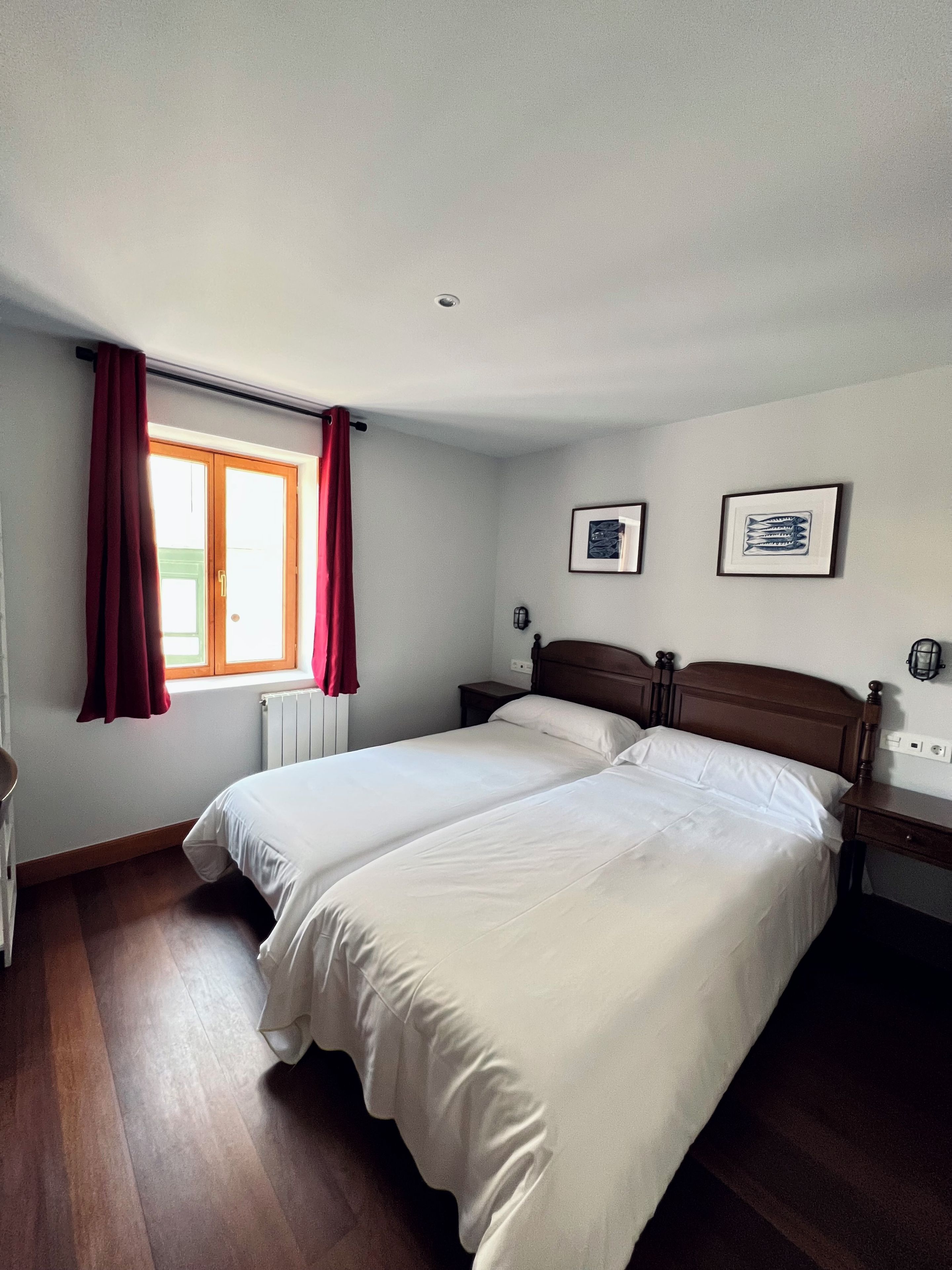 A bedroom with two beds, a window with red curtains, and two framed pictures on the wall.