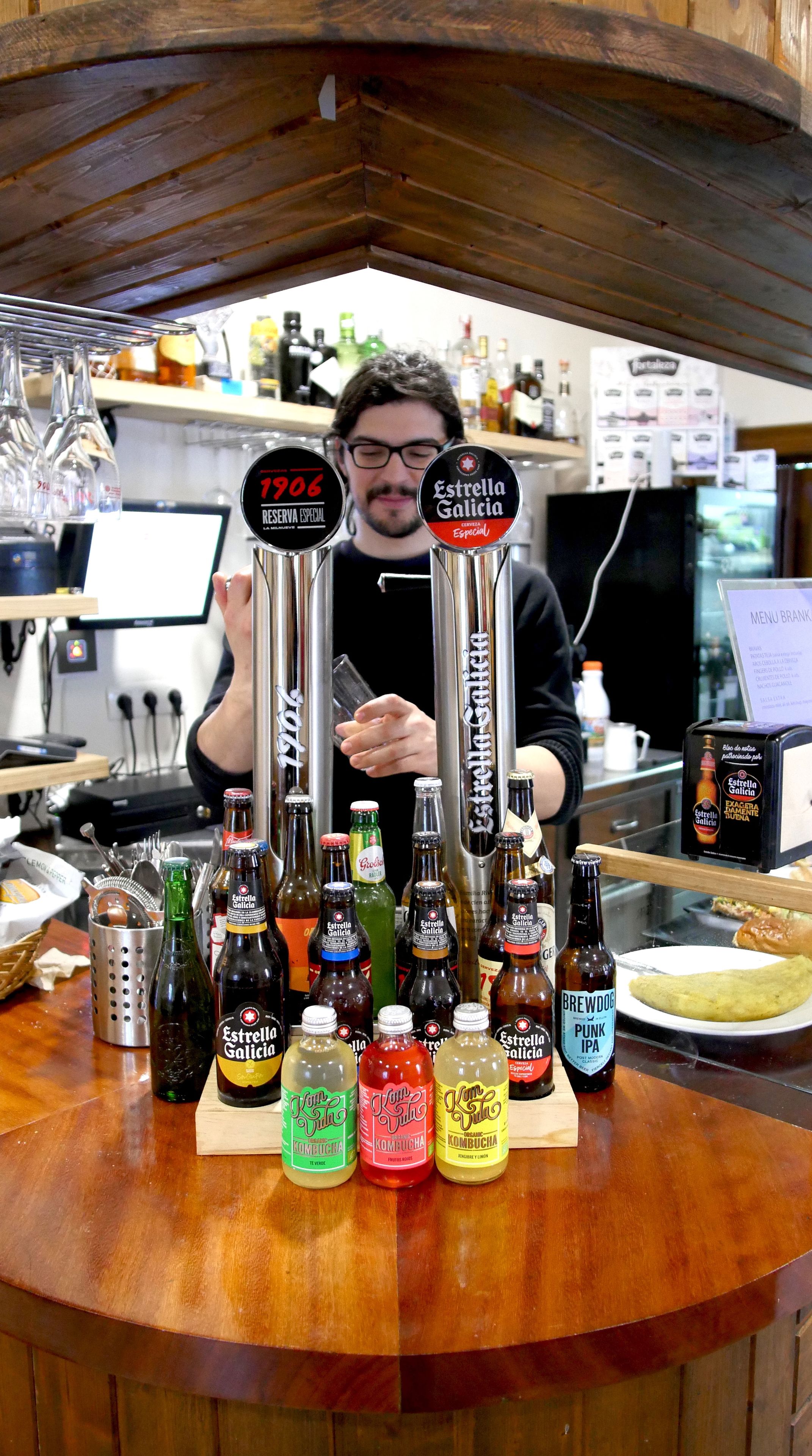 A bar counter with various bottles of Estrella Galicia beer, a bottle of BrewDog Punk IPA, and a bottle of Kombucha. There is a person behind the counter, and a menu sign is visible.