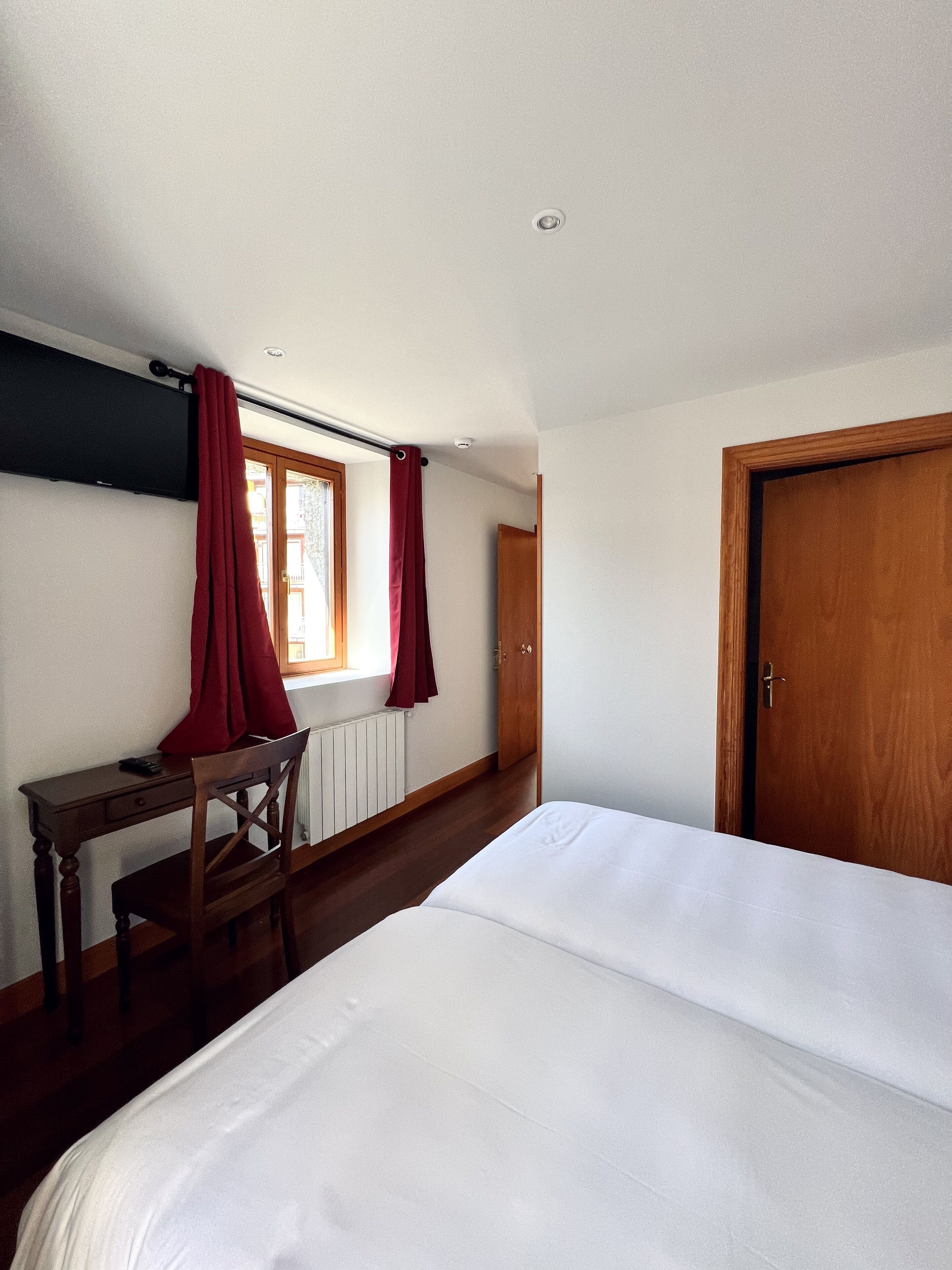A bedroom with a bed, a desk, and a chair. There is a window with curtains, a door, and a radiator.